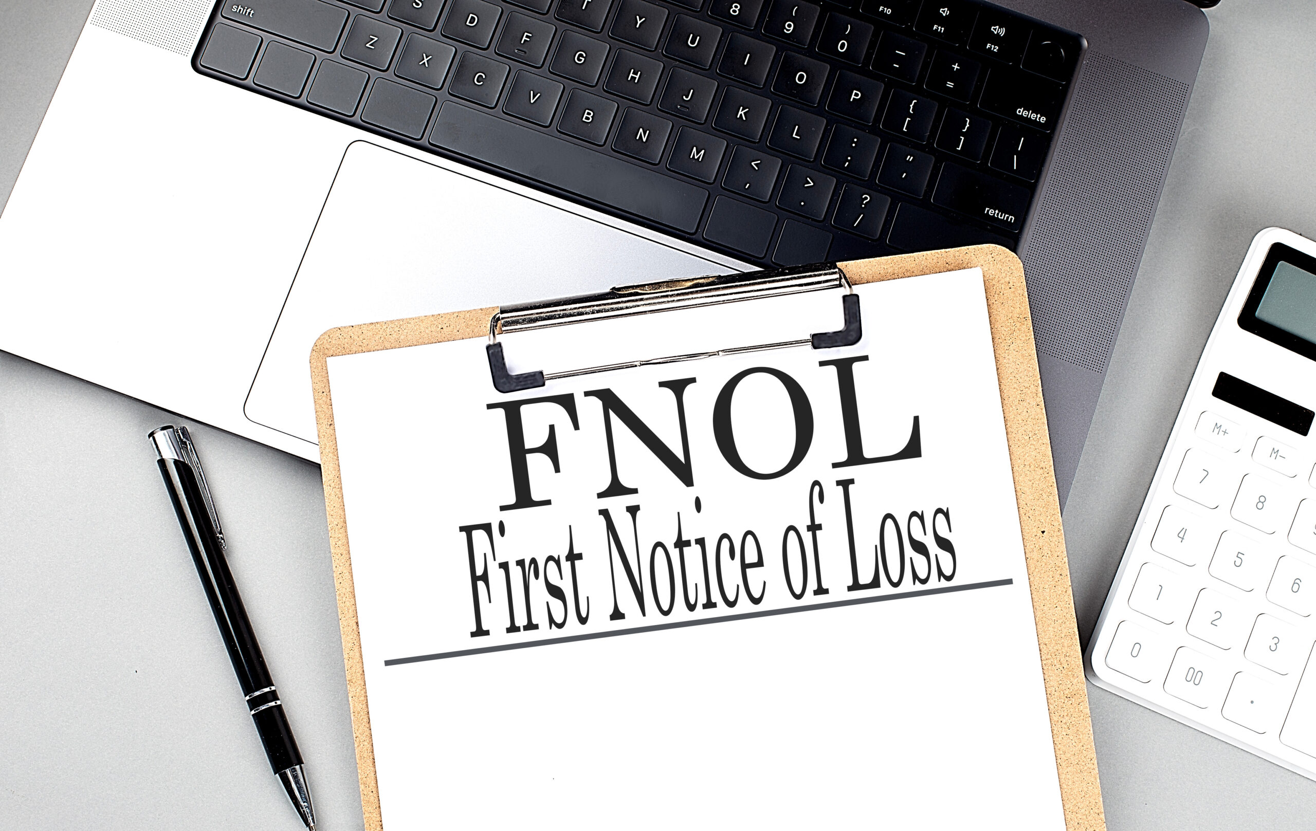 FNOL first notice of loss text on clipboard with laptop, calculator, and pen on grey desk